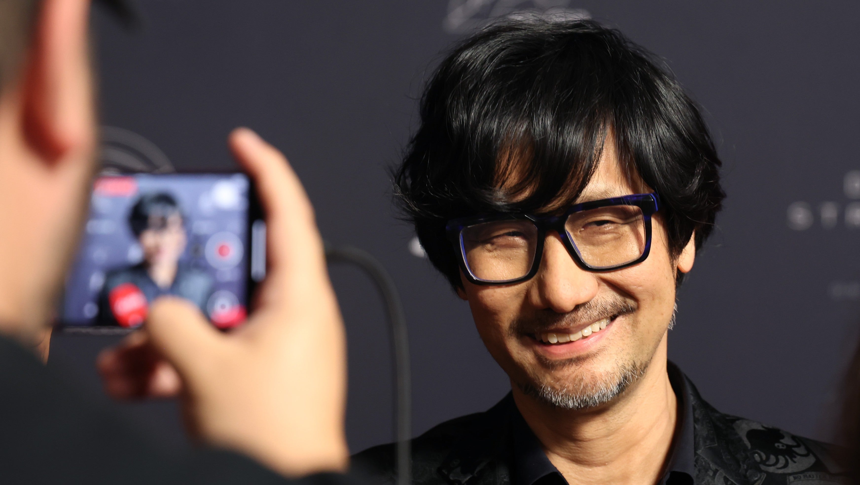 Hideo Kojima acabou de publicar fotos em frente a sede da Sony em Tóquio usando uma camiseta do Joy Division, e o mundo dos games não consegue parar de teorizar. Meses depois de a Sony cancelar Physint, o sucessor espiritual de Metal Gear, os fãs comparam a visita a Johnny Silverhand invadindo a Arasaka Tower em Cyberpunk 2077, e os memes não param.

A história por trás disso é um lembrete cruel de como a criatividade virou algo descartável dentro das grandes corporações dos games. Kojima teria descoberto o cancelamento do projeto por uma simples chamada de Zoom, chegando a temer pelo futuro da Kojima Productions antes de a Xbox salvar o jogo. Essa é a realidade de uma indústria em que decisões de diretoria podem destruir um estúdio da noite para o dia enquanto os desenvolvedores pagam a conta.

Kojima garante que não há mágoas e afirma que ainda ama a Sony após 30 anos, mas o mistério continua. Colaboração secreta, um novo capítulo de Death Stranding ou apenas uma camiseta e uma visita amistosa? Seja o que for, a lenda dos games segue fazendo todo mundo especular.