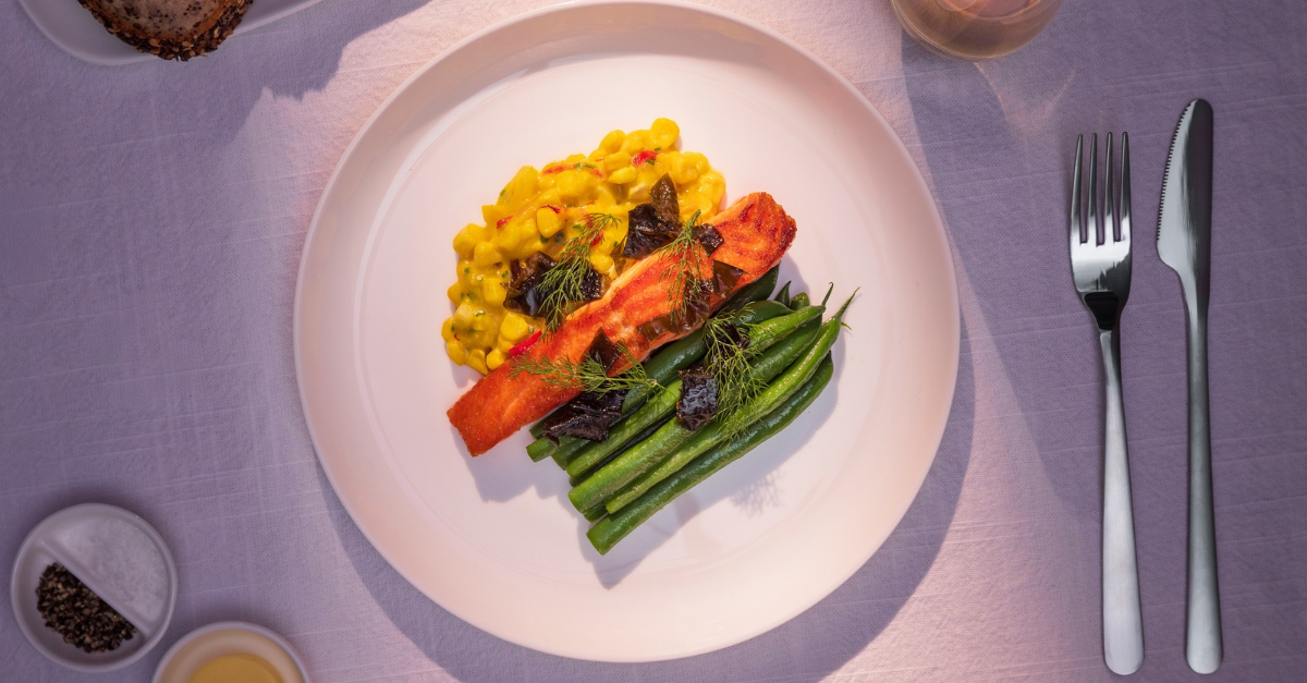 A Air New Zealand entra de vez na disputa das cabines premium com um novo cardápio de longa distância assinado pelo chef neozelandês Josh Emett, que conquistou estrelas Michelin em restaurantes de Londres, Nova York e Los Angeles. A seleção aposta em ingredientes locais, do cordeiro neozelandês ao salmão Alpine e ao cervo selvagem de Fiordland, além de sobremesas com kiwi, biscoito Anzac e outros símbolos do país.

A introdução é gradual a partir de outubro de 2026 nas cabines Premium Economy, Business Premier e Business Premier Luxe dos voos de longa distância que partem de Auckland, com rotas selecionadas saindo da América do Norte. Emett desenvolve as receitas ao lado das equipes de gastronomia da companhia e de produtores locais, considerando que pressão atmosférica, umidade e ruído alteram a percepção dos sabores durante o voo.

O movimento mostra que as aéreas voltaram a competir pelo paladar e pela origem dos ingredientes, apostando que uma refeição memorável a 10 mil metros vale mais do que mais alguns centímetros de espaço para as pernas. Para quem voa a Auckland, cidade erguida sobre mais de 50 vulcões extintos e cercada por duas baías, agora existe mais um motivo para aguardar o próprio voo.